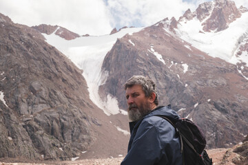 Portrait of a mature bearded man with gray hair and backpack, standing in front of dramatic rocky mountains and glacier. 