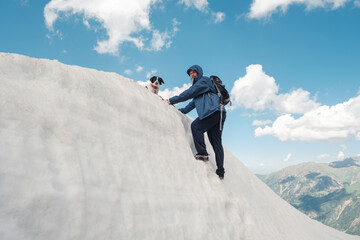 Man in a blue jacket and backpack climbing a steep snow-covered slope with his dog at the top, under a bright blue sky with scattered clouds. 