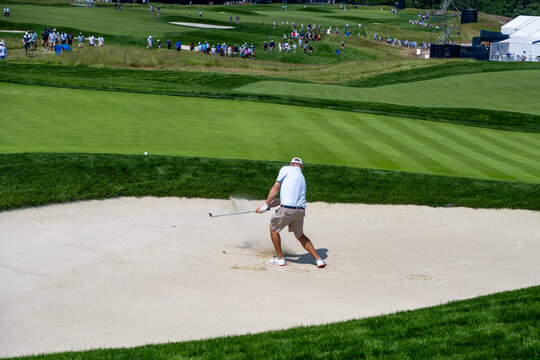  A golfer, captured in mid-swing, hits a golf ball from a sand bunker, sending sand flying. The dynamic shot is set against a lush green golf course. - Powered by Adobe