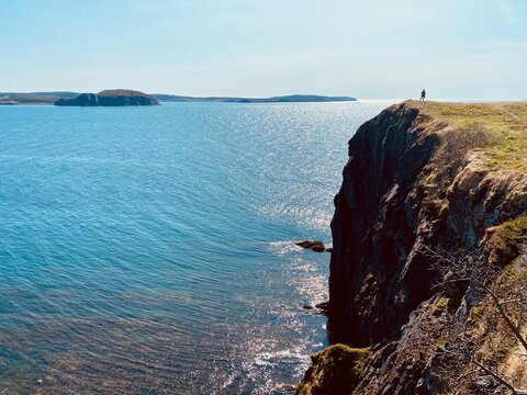 A person on the edge of a cliff admiring the sparkling atlantic ocean, enjoying the skerwink trail, outside Trinity, Newfoundland, Canada