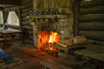 A large stone fireplace with a roaring fire dominates a rustic log cabin interior. Benches flank...