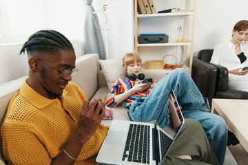 Young man working on laptop while friends and coworkers socialize in casual workplace, highlighting...