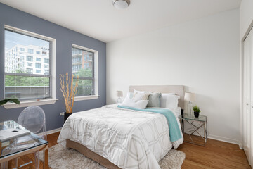 A bedroom with white and blue walls, cozy bedding in a naturally lit room, and a small glass desk and clear plastic chair.