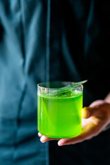 Close-up of a hand holding a glass of bright green liquid against a dark background. Vivid drink with bold color contrast and modern, minimalistic composition