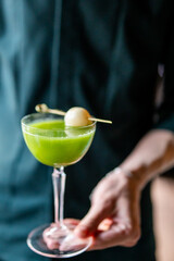 Close-up of a hand holding a cocktail glass with a green drink, garnished with a pickled onion on a skewer, isolated against a blurred background. Focus on the vibrant beverage