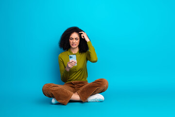 Young Woman With Curly Hair Sitting Against Blue Background Using Smartphone With Confused...