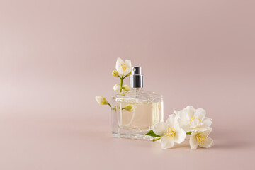 A beautiful glass perfume bottle stands on a beige stone background with delicate white flowers and a sprig of jasmine. A blank bottle mockup. space.