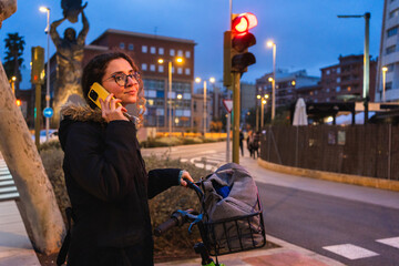 Young woman using mobile phone while waiting with her bicycle at red traffic light in urban environment at dusk