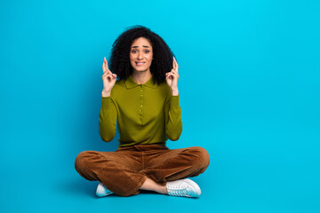 Young woman seated casually on a blue background crossing fingers in hope wearing a stylish khaki...