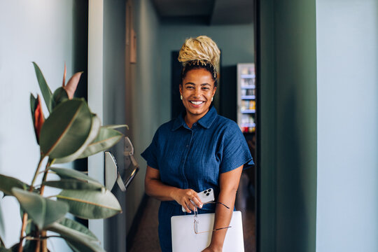 Smiling woman standing in office hallway holding laptop and smartphone