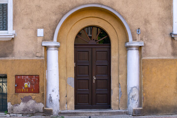 Wooden entrance door set in a yellow wall with a rounded arch and two columns. Visible signs of...