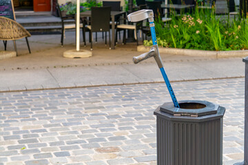 Discarded crutch sticking out of a trash bin on a city sidewalk, with a café and greenery in the...