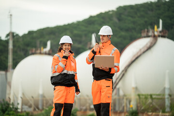 Two engineers wearing safety gear are working at an industrial site near spherical gas storage tanks. Using laptop and walkie talkie to coordinating field operations in a petrochemical environment