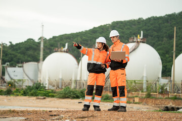 Two engineers in safety gear talking work near spherical gas storage tanks. woman is pointing forward while the man checks a laptop, indicating active discussion and site coordination in an industrial