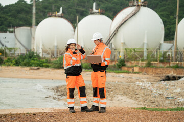 Two engineers wearing safety gear are working at an industrial site near spherical gas storage tanks. Using laptop and walkie talkie to coordinating field operations in a petrochemical environment