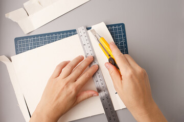 person cutting white cardboard with yellow utility knife and metal ruler on blue grid mat ideal for...