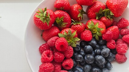 Fresh strawberries, raspberries, and blueberries arranged in a white bowl under natural light.