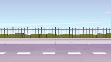 A low-angle view of an empty roadside featuring a fence and a row of green bushes.  The scene is set under a light blue sky, suggesting a calm and peaceful day.