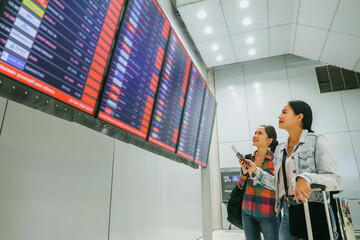Two female asian passengers who were friends holding tickets for an international trip stood in the departure terminal and looked at the departure board with interest to check their own flights.