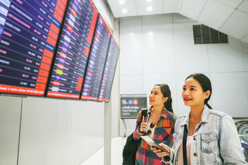 Two female asian passengers who were friends holding tickets for an international trip stood in the departure terminal and looked at the departure board with interest to check their own flights.