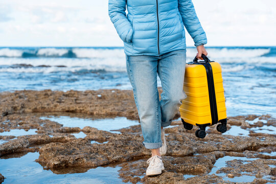 Person walks along rocky shore holding a yellow suitcase, ocean waves gently crashing on the beach during bright daylight hours - Powered by Adobe