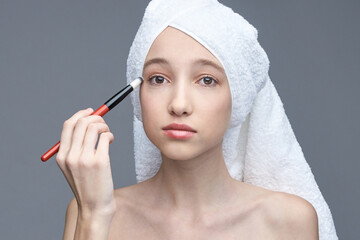 Young caucasian female applying makeup with brush against gray background.