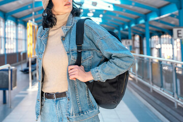 Young woman in denim jacket with backpack stands at a vibrant train station, surrounded by bright...