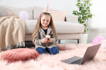 Birthday celebration. Little girl with cupcake having video call via laptop at home
