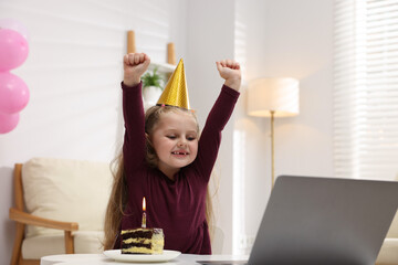 Birthday celebration. Little girl with piece of cake having video call via laptop at table indoors