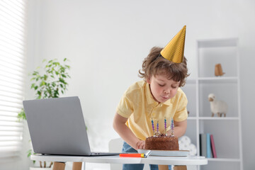 Birthday celebration. Little boy blowing candles on cake while having video call via laptop at...