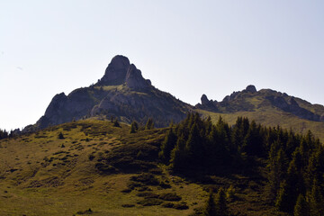 Majestic mountain peaks rise above rolling green hills and pine forests in this scenic landscape of...