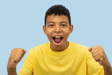 Cheerful young brown skinned Latino man wearing a yellow shirt celebrating with both hands on a light blue background with copyspace