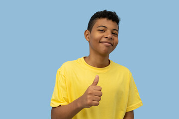Cheerful dark skinned Latino teenager wearing a yellow shirt making a thumbs-up gesture on a light blue background with copyspace