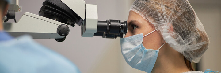 woman wearing surgical mask and hairnet using operating microscope in medical operating room,...