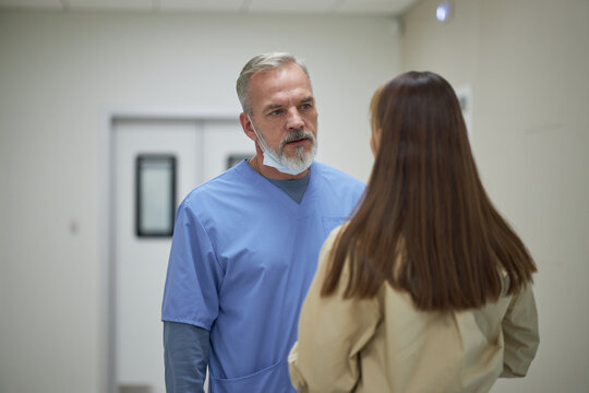 Middle aged male doctor wearing scrubs talking to woman in hospital corridor, both standing near operating room doors, discussing medical situation