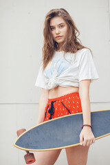 Young stylish woman in red shorts holding a longboard, standing against a white wall. Summer urban fashion vibe.