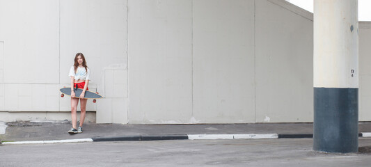 Minimalist shot of a young woman with skateboard standing alone against a blank concrete wall. Urban youth style.