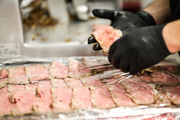 Close-up of hands in black gloves packaging sliced roast beef in plastic wrap, representing food preparation and hygiene in a professional kitchen.