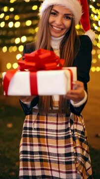 Holiday Gift Giver: A joyous figure dressed in a festive Santa hat offers a beautifully wrapped present adorned with a bright red bow, illuminated by the warm glow of string lights.