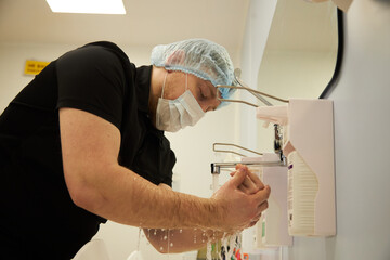 Middle aged man wearing surgical mask and hair cover washing hands at medical sink in operating room preparing for surgery, focusing on thorough hand hygiene before procedure