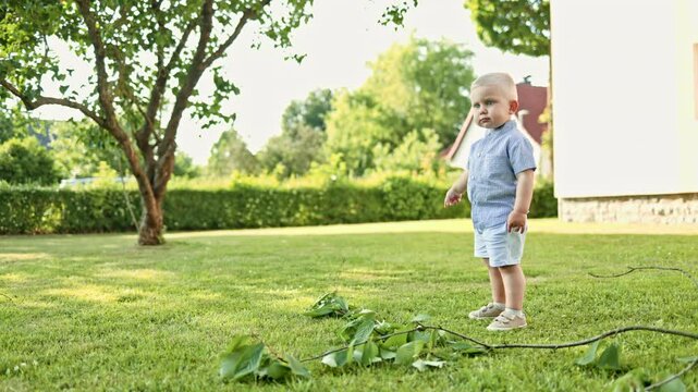 A white-haired 2-year-old boy in shorts and a shirt runs through the grass on a summer day. A boy in a stylish outfit on a walk in the park. The baby is running. self-development ditini.