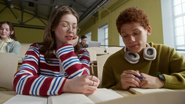 Medium shot of diverse zoomers sitting at desk discussing notes during group lecture in spacious auditorium