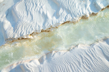 Hot mineral water stream running over textured white travertine surface in Pamukkale, Turkey. Close-up view showing natural erosion patterns and subtle color variations under soft daylight.