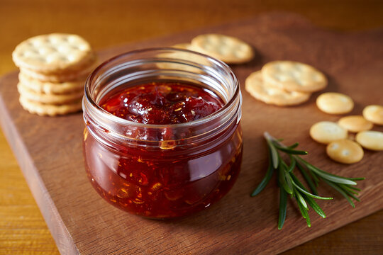 Glass jar filled with homemade red pepper jelly served with crackers and fresh rosemary sprig - Powered by Adobe