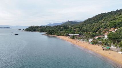 Aerial drone footage of Praia Grande in Ilhabela, SP, Brazil, with light sand, Atlantic forest in the background, and clear ocean water under a sunny summer sky