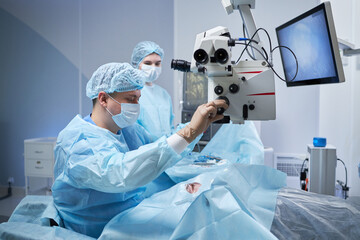 middle aged man performing surgery using operating microscope while man assisting in modern...