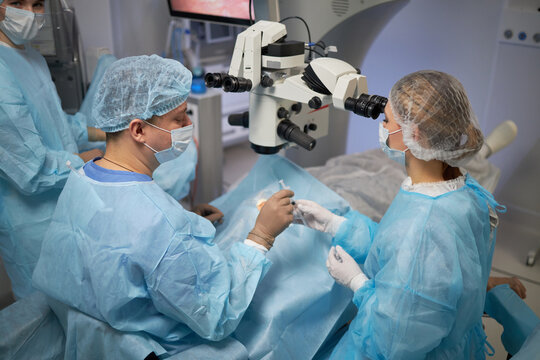 middle aged man and woman performing surgery in operating room using surgical microscope, medical staff assisting, patient lying on operating table