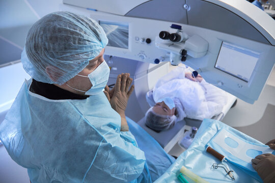 Middle aged male surgeon preparing for eye surgery on woman lying under ophthalmic laser system in modern operating room with medical instruments visible