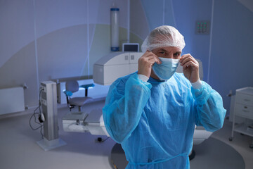 Fototapeta premium middle aged man wearing surgical gown and cap adjusting face mask in operating room preparing for medical procedure with advanced equipment visible in background