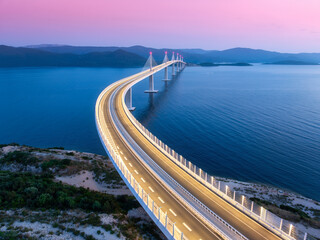 Aerial view of modern Peljesac bridge and blue sea at night. Croatia. Bridge with illumination, mountains, reflection and pink sky at sunset. Top drone panoramic view of road, street lights at dusk